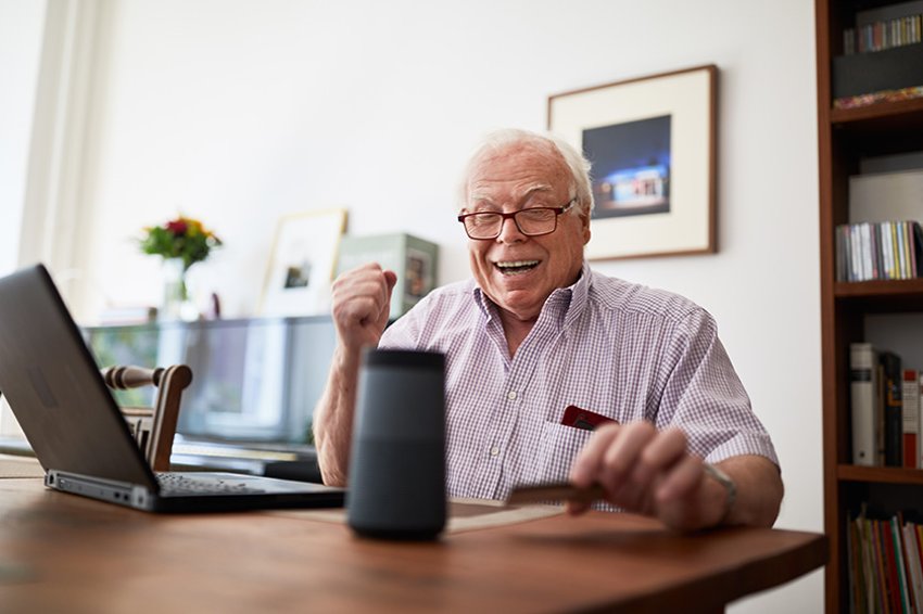 Älterer Mann beim Online-Shopping mit Kreditkarte, Laptop und Smart-Speaker-Gerät.